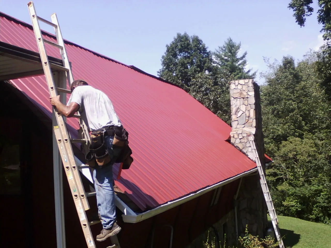 Roofer on a ladder beside a red metal roof and stone chimney — Soffit & Fascia Repair by Roofing Brothers in Haymarket